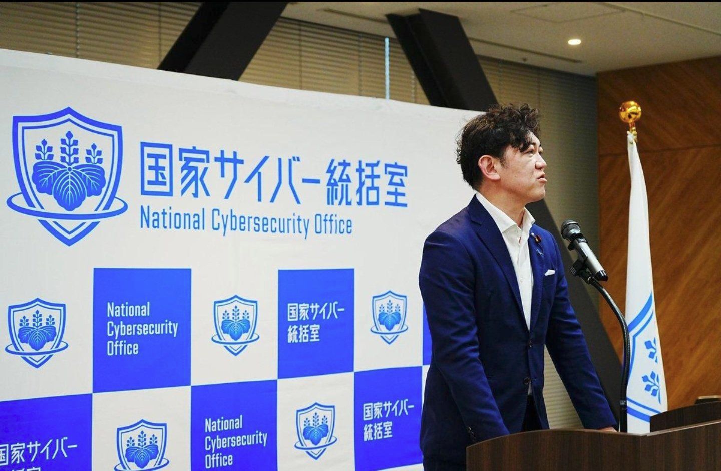 Un funcionario habla desde un atril frente al panel de la Oficina Nacional de Ciberseguridad de Japón