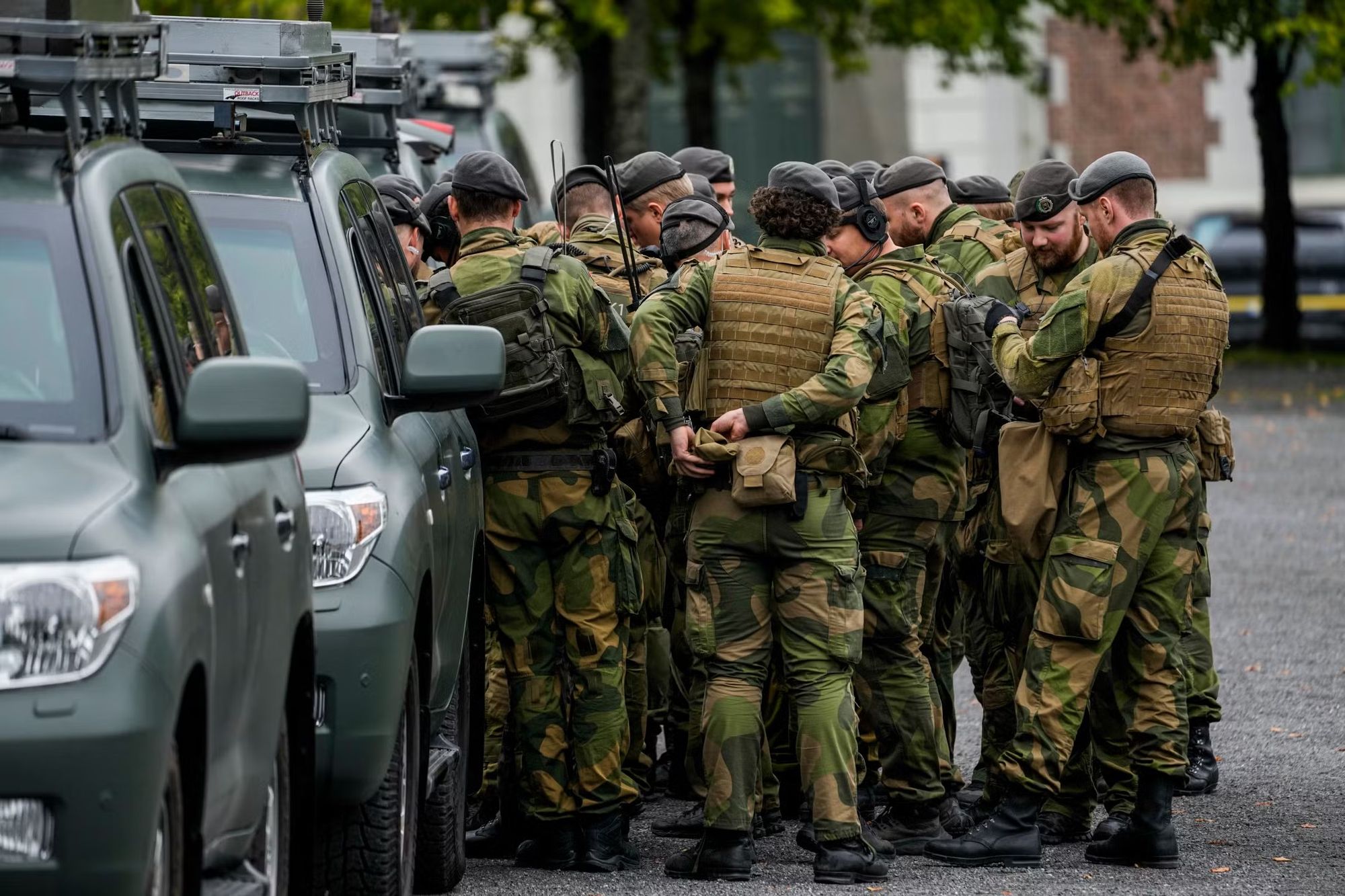 Grupo de soldados noruegos con uniforme de camuflaje, boinas y chalecos tácticos reunidos junto a una fila de camionetas verdes