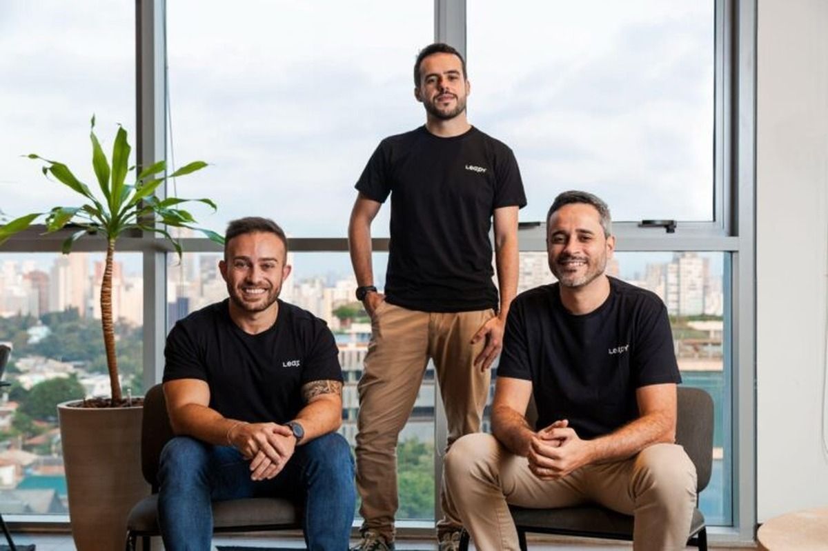 Los tres cofundadores de Leapy, con remeras negras y pantalones claros, sentados y de pie en una oficina con ventanales y vista a una ciudad