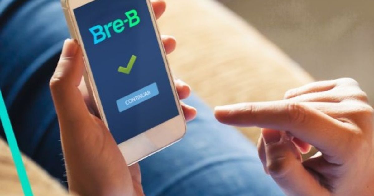 Manos sosteniendo un celular con el logo de Bre-B y una marca de verificación sobre un botón Continuar, mientras un dedo de la otra mano se acerca a la pantalla