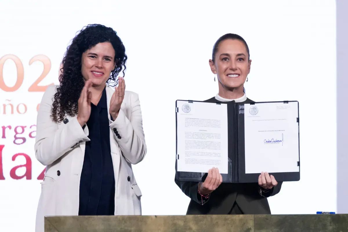 La presidenta Claudia Sheinbaum muestra el decreto firmado, junto a una funcionaria que aplaude, en la conferencia matutina