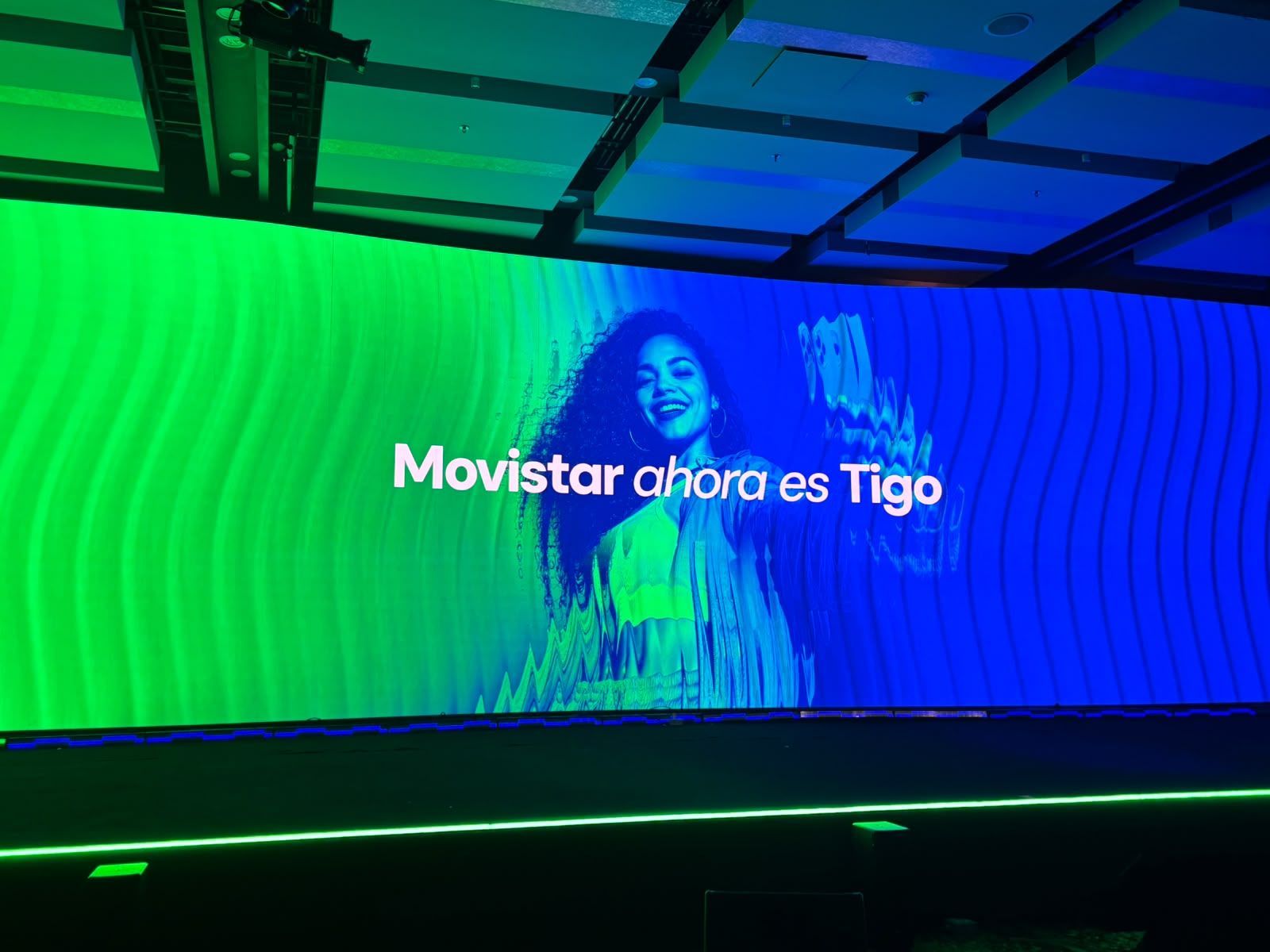 Pantalla gigante en un auditorio con el mensaje "Movistar ahora es Tigo" y la imagen de una mujer sonriente sobre un fondo verde y azul
