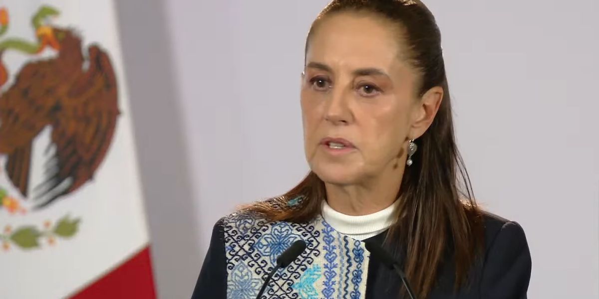 La presidenta Claudia Sheinbaum habla frente a dos micrófonos, junto a la bandera de México, durante su conferencia matutina