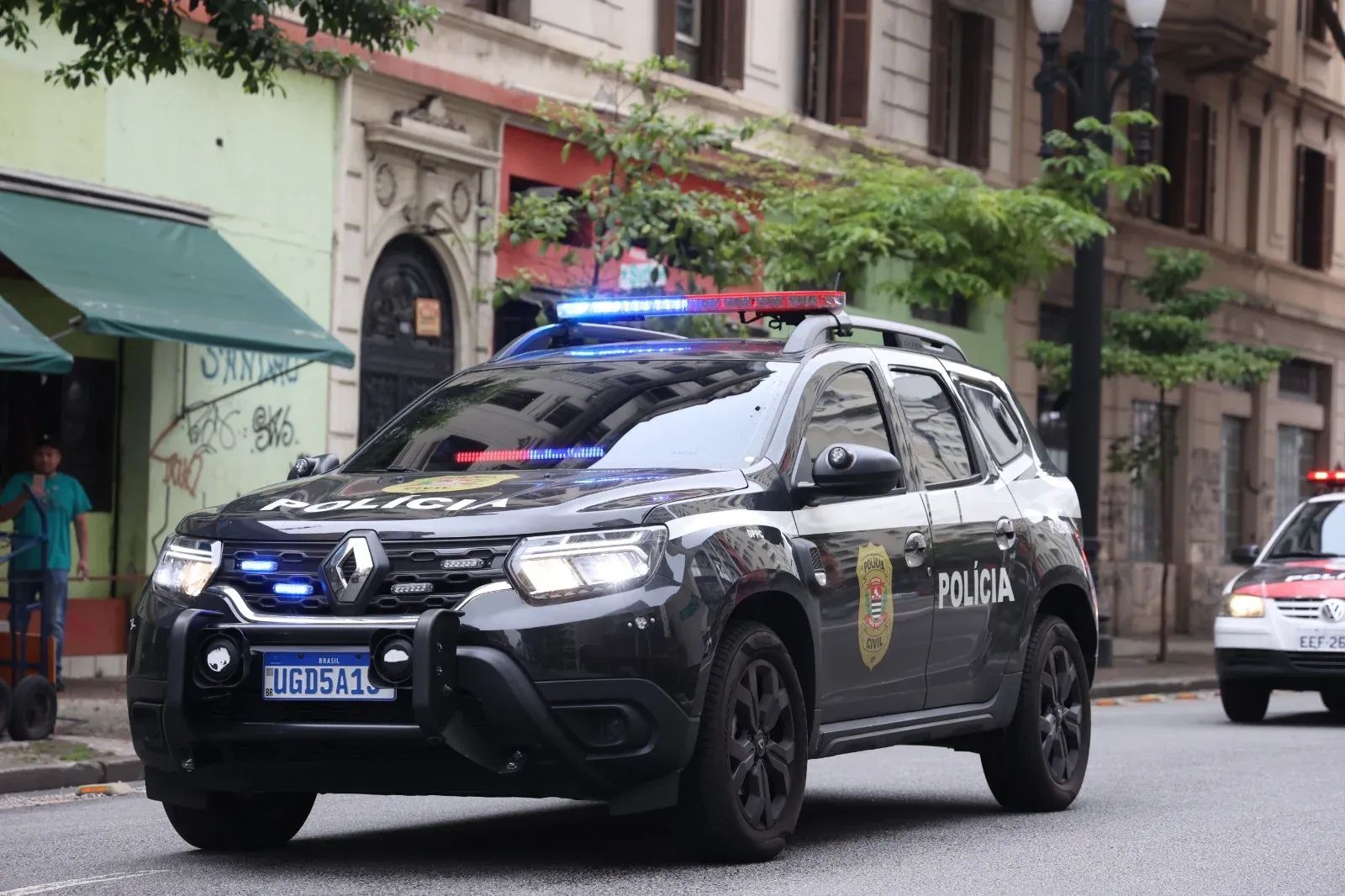 Patrulla negra de la Policía Civil con luces azules y rojas encendidas sobre el techo, avanzando por una calle de la ciudad. Se lee POLÍCIA en el capó y en la puerta, y la placa azul dice UGD5A15. Detrás, otro vehículo blanco y rojo.