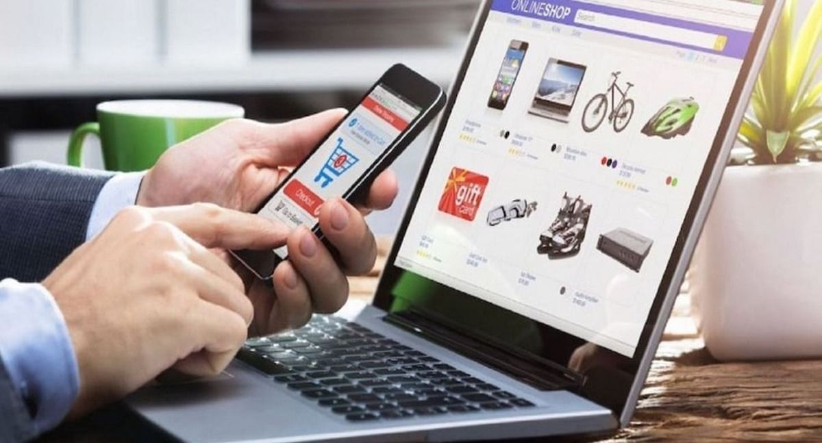 Dos manos sostienen un celular con un carrito de compras en pantalla, frente a una laptop que muestra una tienda en línea