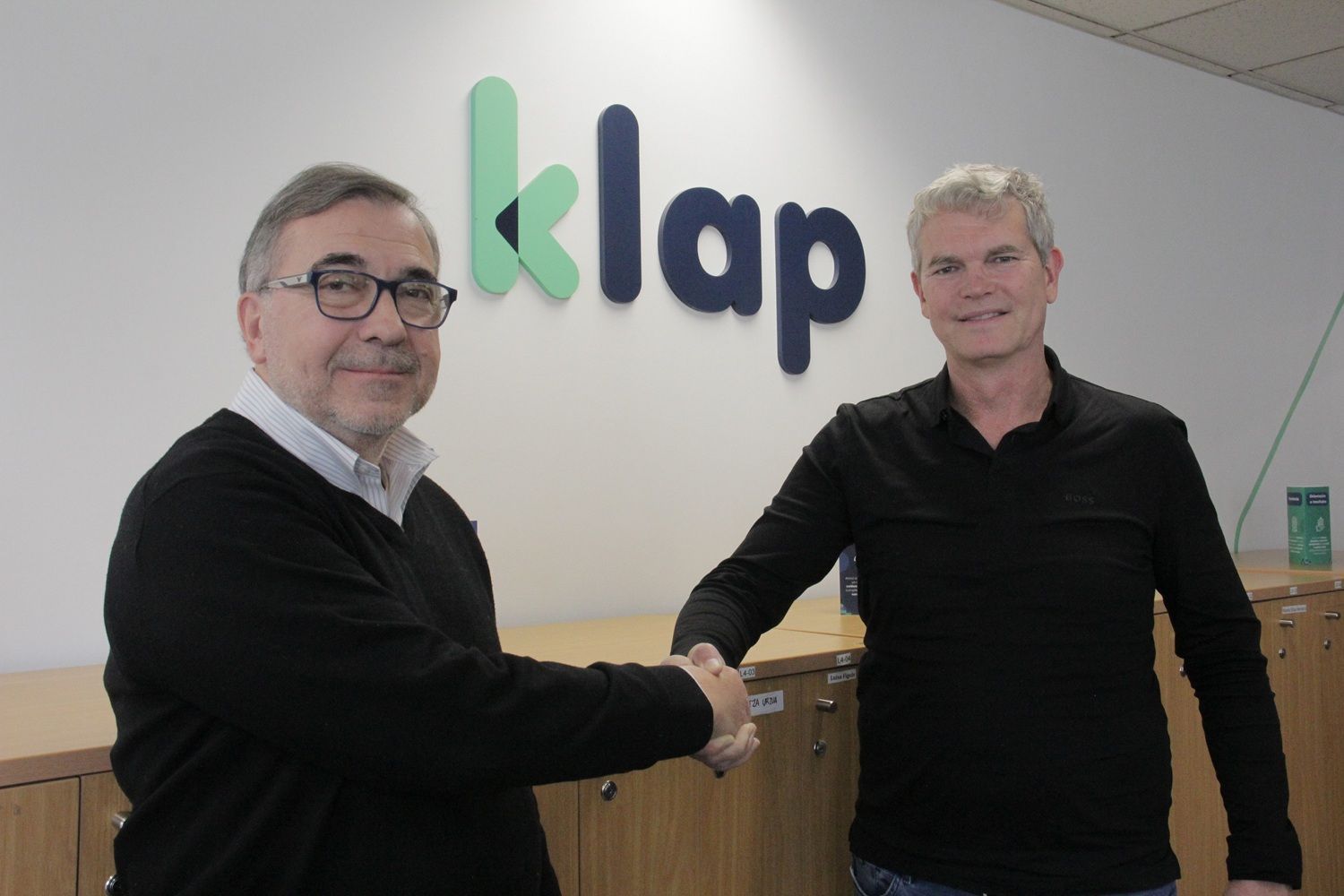 Dos hombres se dan la mano frente a una pared blanca con el logotipo verde y azul de Klap, en las oficinas de la empresa