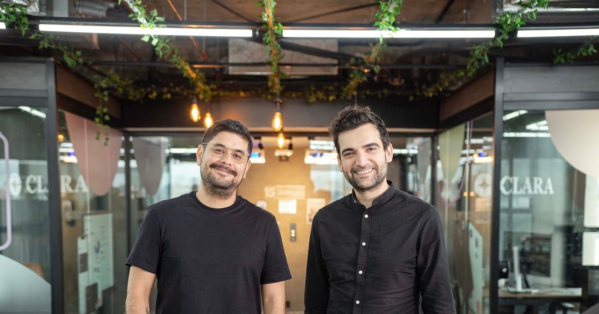 Dos hombres sonrientes con camisas oscuras posan de frente en una oficina con paredes de vidrio, plantas colgantes y el logo de Clara en los cristales