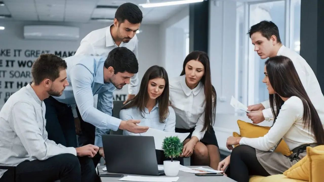 Seis jóvenes profesionales reunidos alrededor de una computadora portátil en una oficina, mirando unos papeles