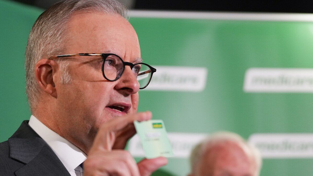 Anthony Albanese, de anteojos, muestra una tarjeta de Medicare frente a un fondo verde con el logo del sistema