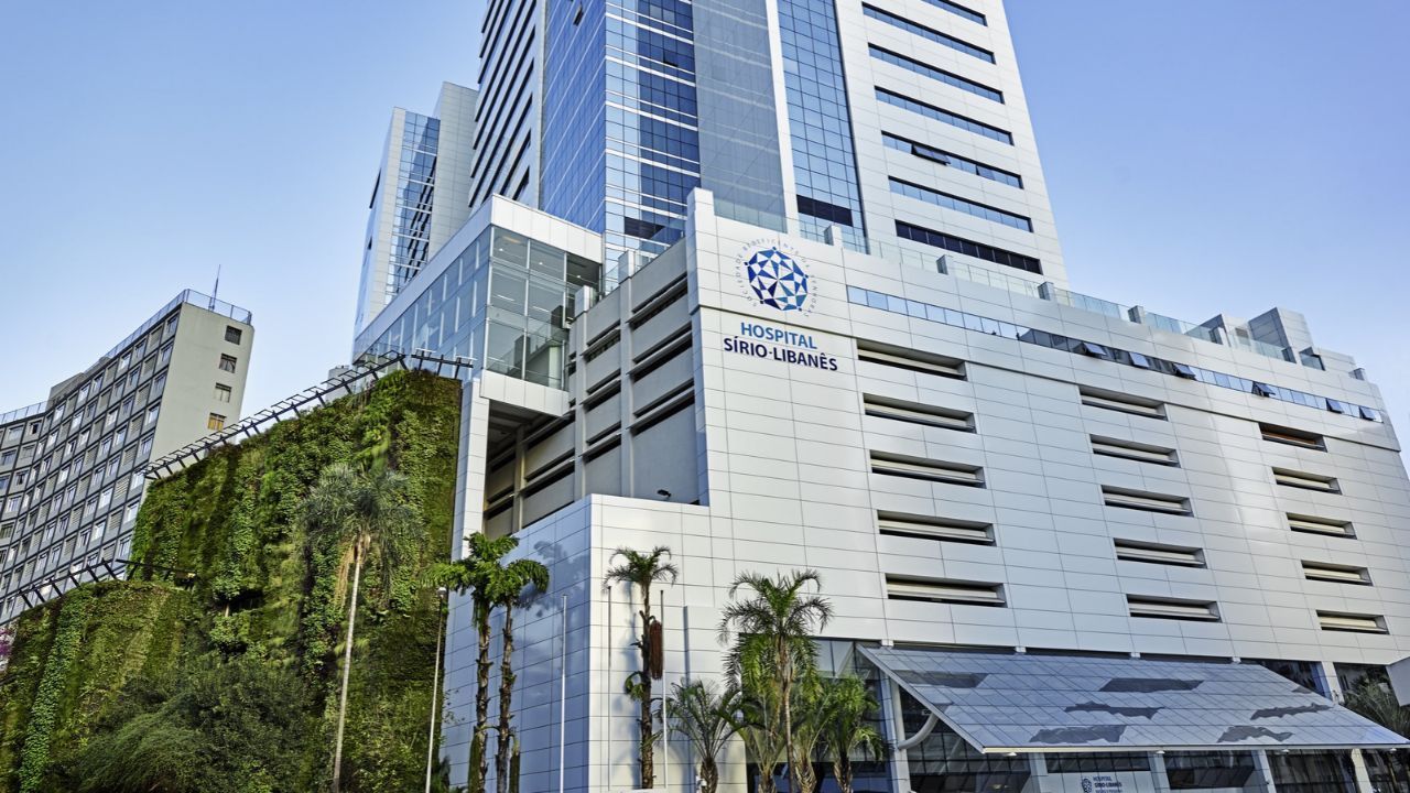 Fachada del edificio del Hospital Sírio-Libanês, con su logotipo y nombre en la pared blanca, una torre vidriada detrás y palmeras en la entrada