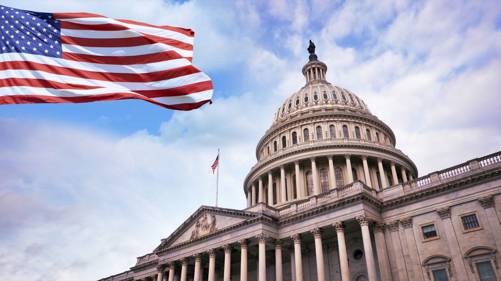 La cúpula del Capitolio de Estados Unidos bajo un cielo con nubes, con la bandera estadounidense flameando en primer plano