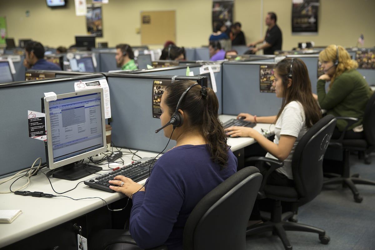 Oficina con cubículos de call center donde varias personas trabajan frente a computadoras con auriculares con micrófono. En primer plano, una mujer de camisa azul marino mira la pantalla de su monitor, que muestra una planilla o formulario con texto,