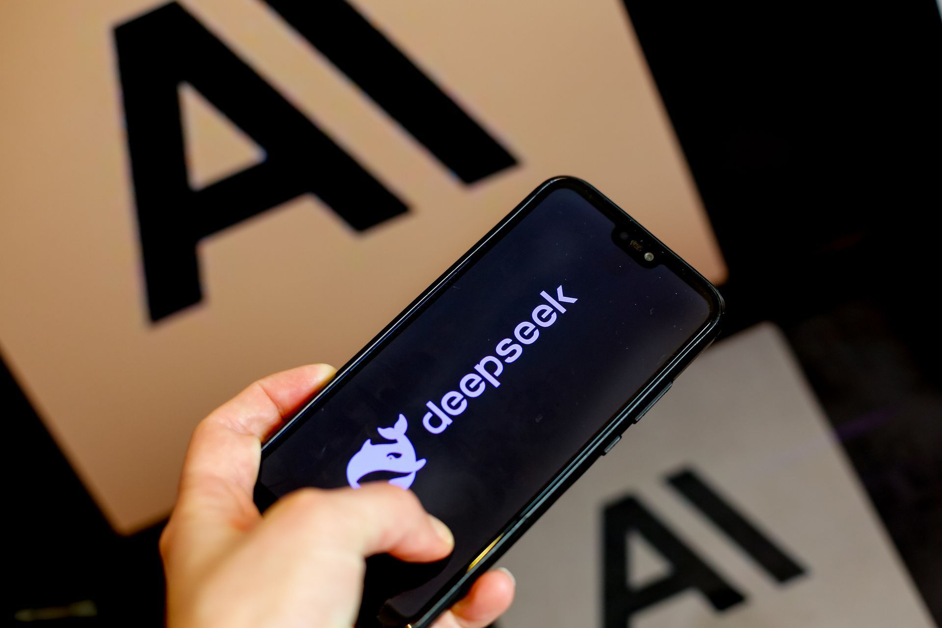 Una mano sostiene un celular con el logo de DeepSeek en la pantalla, frente a carteles con las letras AI