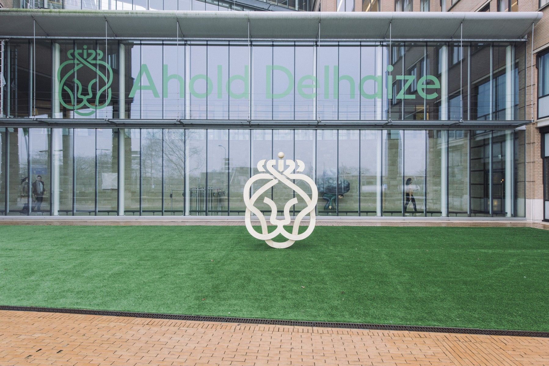 Fachada vidriada de la sede de Ahold Delhaize en Zaandam, Países Bajos, con el nombre y el logo del león del grupo frente a un patio de césped