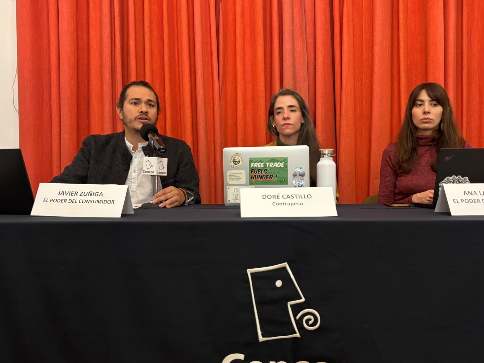 Javier Zúñiga, de El Poder del Consumidor, habla en una conferencia de prensa junto a Doré Castillo y Ana Larrañaga, frente a un telón naranja