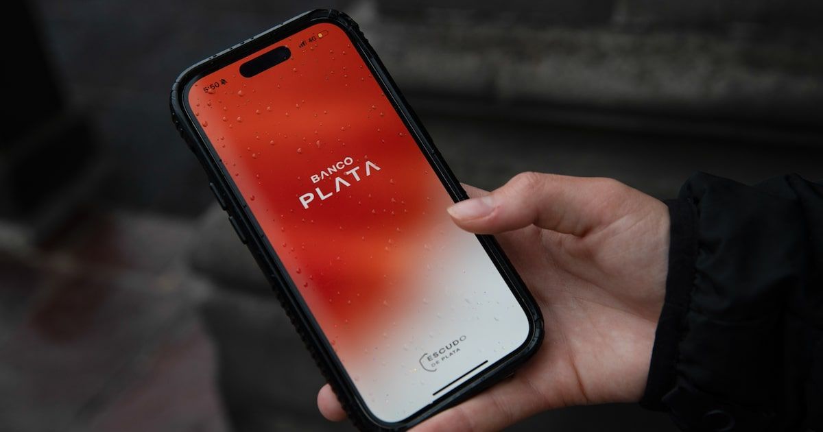 Una mano sostiene un celular con la pantalla de inicio de la app de Banco Plata, en rojo