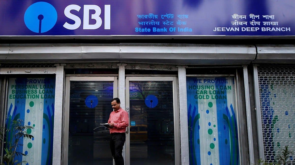 Fachada de una sucursal de State Bank of India con el cartel azul del banco y un hombre leyendo papeles frente a la puerta