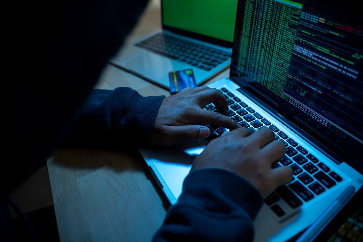 Persona encapuchada escribe en una computadora portátil con líneas de código en pantalla y una tarjeta de crédito sobre la mesa