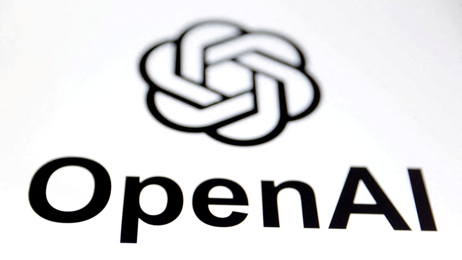 Logotipo de OpenAI en negro sobre fondo blanco, desenfocado: arriba, un símbolo formado por cinco óvalos entrelazados en forma de flor o nudo, y debajo, la palabra OpenAI en letras de palo gruesas.