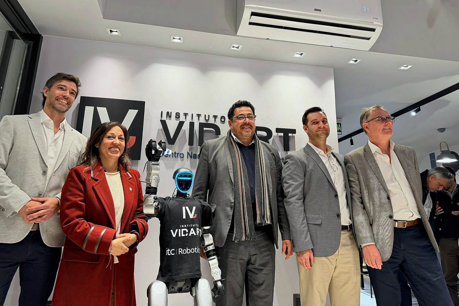 Cinco personas posan sonrientes frente a un panel blanco con el logo IV y el texto Instituto Vidart. Delante, un robot humanoide con cabeza negra con luz azul y una remera negra que dice Instituto Vidart e itc Robotics. A la izquierda, una mujer de c
