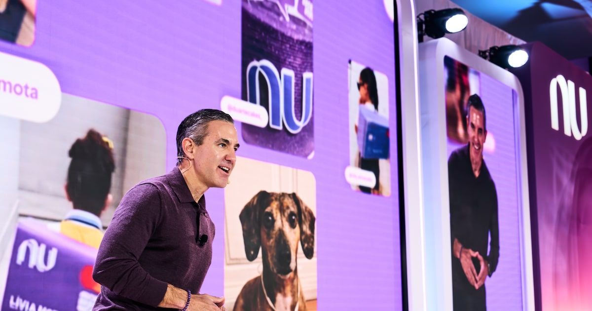 Un hombre de pelo canoso, con camisa violeta oscura y micrófono de solapa, habla en un escenario frente a una pantalla morada con el logo blanco de Nu y varias fotos de personas, entre ellas un perro salchicha marrón.