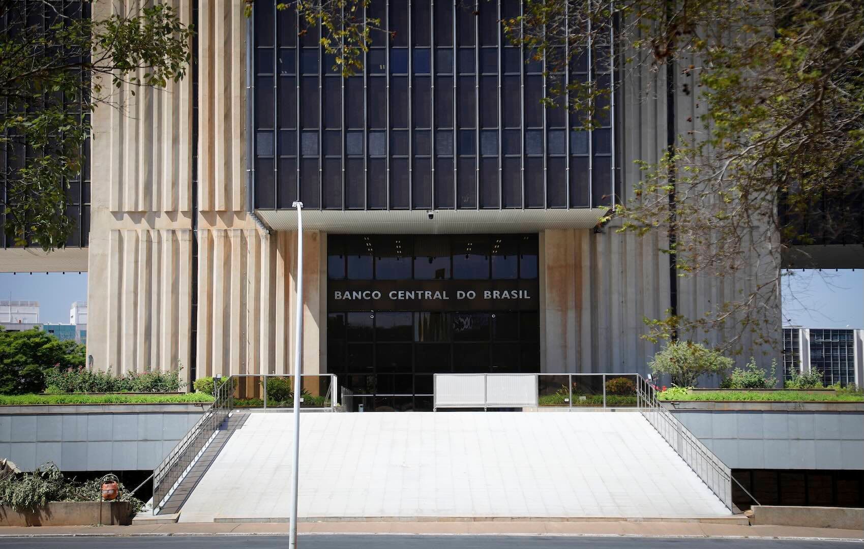 Fachada de un edificio de hormigón de líneas verticales con ventanales oscuros. En el acceso central se lee el cartel BANCO CENTRAL DO BRASIL sobre la puerta. Una escalera ancha y plana con barandas conduce a la entrada, y hay árboles y arbustos a lo