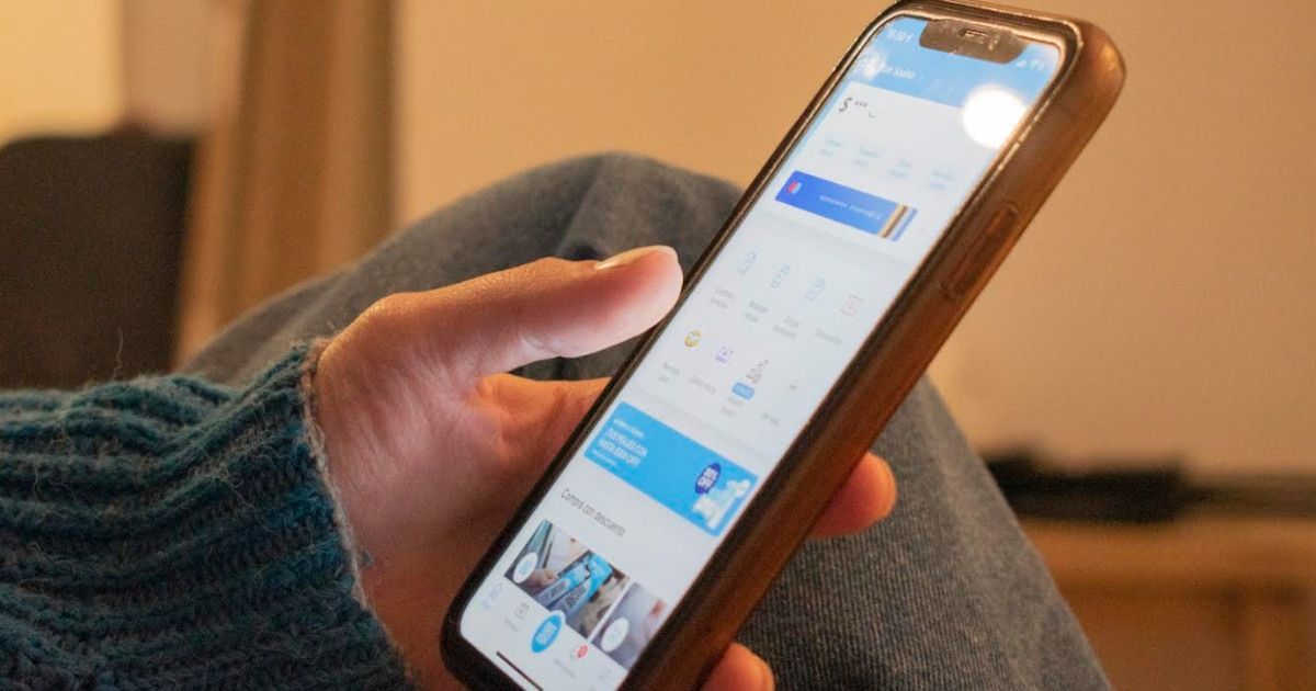 Una persona sostiene un celular con funda marrón y mira la pantalla, donde se ven una app de finanzas con saldo, accesos rápidos y una tarjeta azul de bienvenida. Tiene un suéter verde azulado y el fondo está desenfocado.