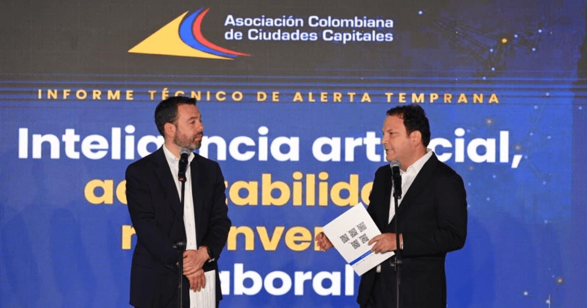 Dos hombres de traje oscuro hablan frente a micrófonos en un escenario. Detrás hay una pantalla azul con el logo de la Asociación Colombiana de Ciudades Capitales y el texto 'Informe técnico de alerta temprana. Inteligencia artificial, accesibilidad'