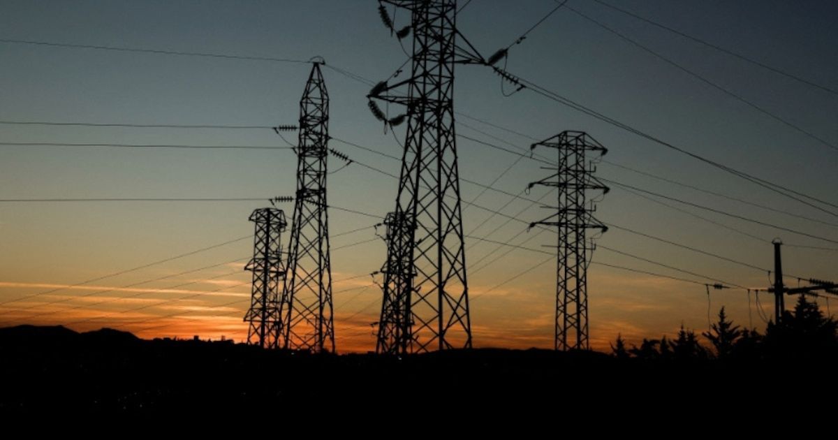 Silueta oscura de varias torres de alta tensión de estructura metálica contra un cielo al atardecer con franjas naranjas y azules; de las torres cuelgan cables y aisladores que cruzan la escena.