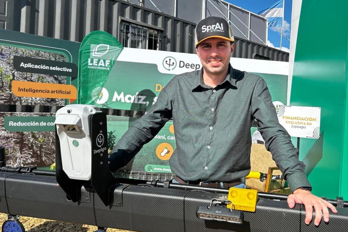 Hombre sonriente con gorra negra que dice SprAI y camisa verde oscura, apoyado sobre un equipo con una cámara blanca marcada DeepAgro. Detrás, paneles con las palabras Aplicación selectiva e Inteligencia artificial.