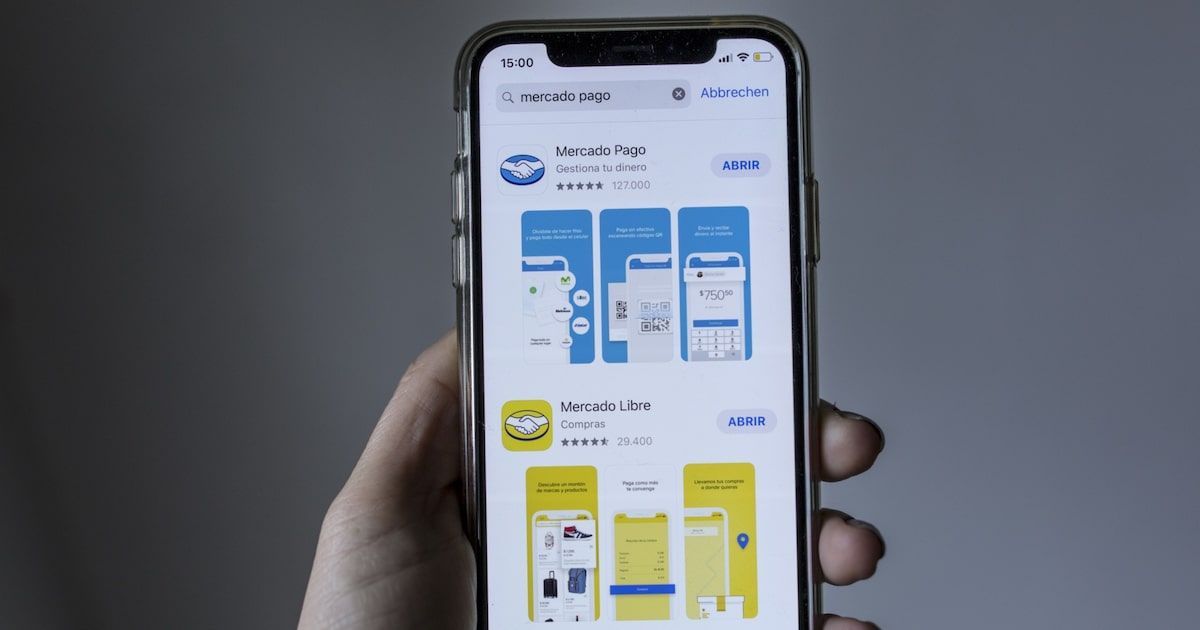 Mano sosteniendo un celular que muestra la App Store con la búsqueda «mercado pago». Aparecen los resultados de las apps Mercado Pago y Mercado Libre, con sus íconos, capturas de pantalla y botones ABRIR.