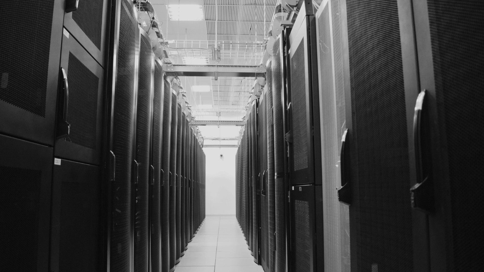 Pasillo central de un centro de datos con filas de racks de servidores negros con paneles perforados a ambos lados, luces de techo encendidas y piso de baldosas claras que se pierde en el fondo. Fotografía en blanco y negro.