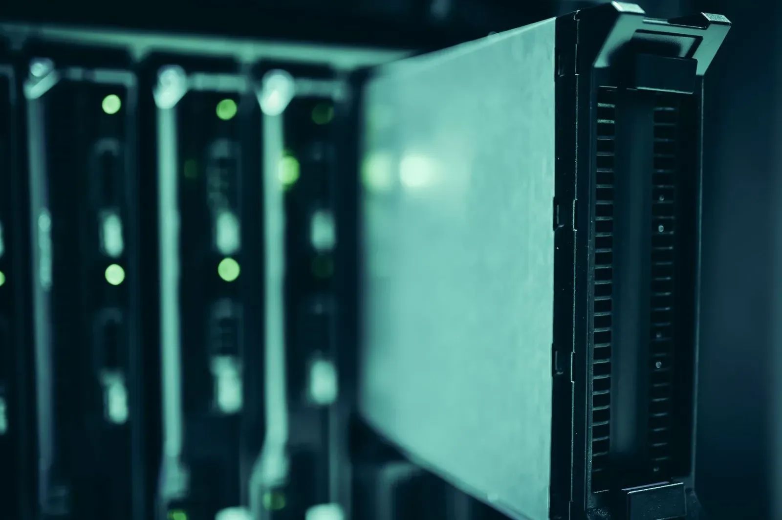 Primer plano de un servidor de centro de datos con panel frontal oscuro y superficie verde azulada, con rejillas de ventilación en el lateral derecho. Detrás, en desenfoque, se ven otros servidores en un rack con pequeñas luces verdes encendidas.