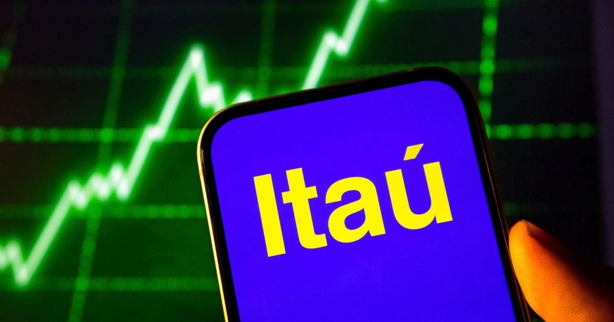 Celular sostenido con una mano, con la pantalla azul que muestra el logotipo de Itaú en letras amarillas. Detrás, un gráfico de líneas verdes luminosas en ascenso sobre fondo oscuro.