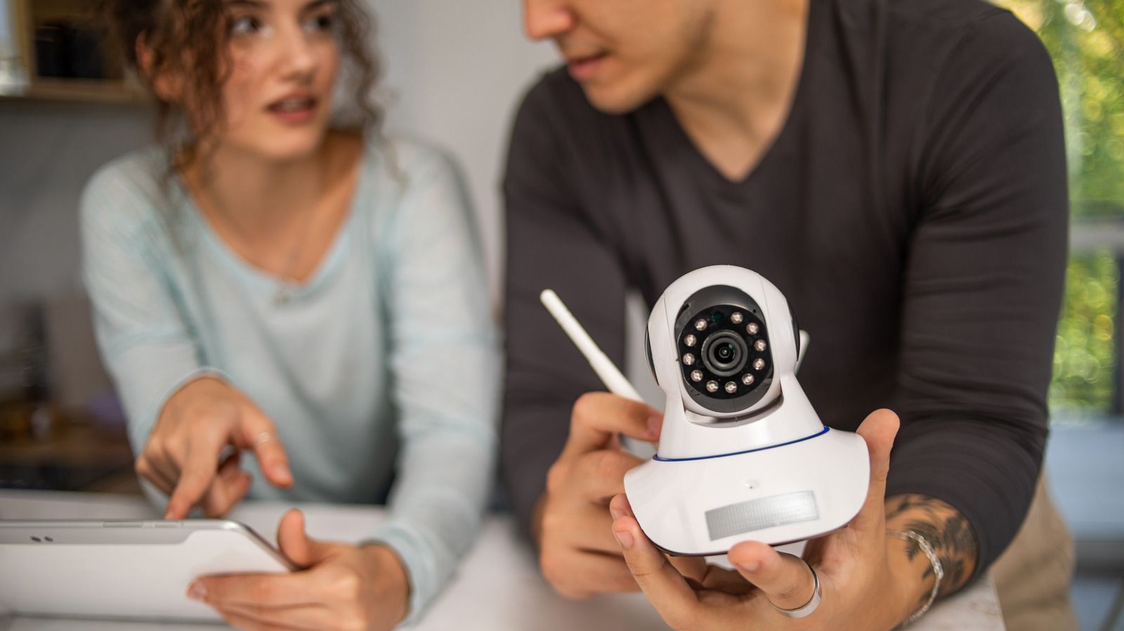 Una pareja en una cocina mira una cámara de seguridad con wifi que sostiene el hombre, junto a una tableta; imagen ilustrativa